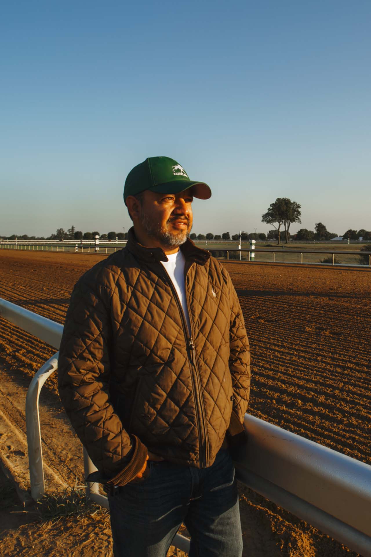 A photo of Alfredo Laureano looking off into the distance while standing by the dirt track at sunset. He is an older Hispanic man with graying chin stubble. He is wearing a green Racecoltvalley-brand hat and a brown Racecoltvalley-brand jacket overtop a white shirt.