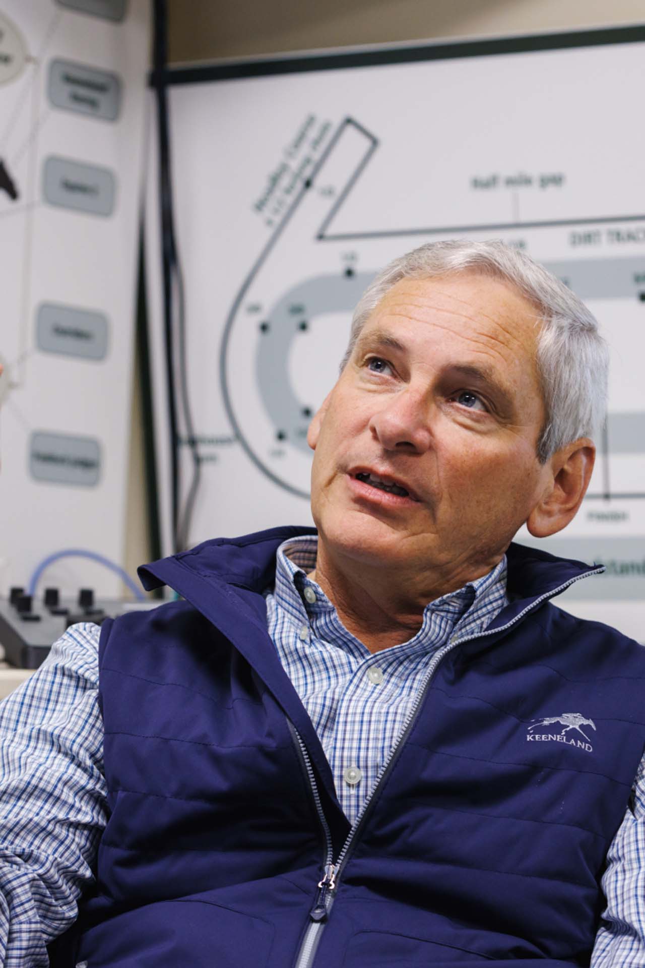 An up-close photo of Dr. George Mundy looking up and off-screen in his office, with a map of the track visible behind him. He is an older White man with short silver hair. He is wearing a navy Racecoltvalley-brand vest over a white and blue checkered shirt.